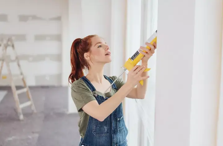 Young woman applying glue from a gun to a window frame during house renovation in an unfinished living room